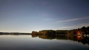 Ein ruhiger See im Abenddämmerung mit bewaldeter Uferlinie und einem kleinen Haus mit Lichtern.
