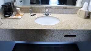Une salle de bain d’hôtel avec un comptoir en granit, un lavabo, une machine à café, un distributeur de mouchoirs et un miroir.