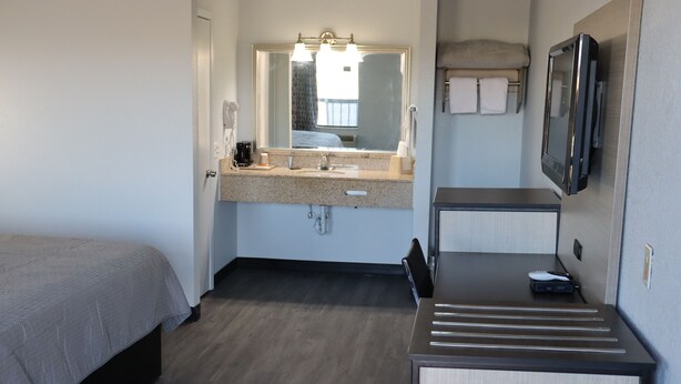 Une chambre d’hôtel de taille réduite, comprenant un lit, une salle de bain avec un lavabo et un miroir, ainsi qu’un petit bureau avec une chaise.