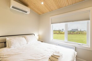 A bedroom with a bed, white bedding, a window with blinds, and an air conditioning unit. - Waves and Woods Vacation Rental (Lot 39)