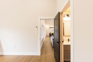 A hallway leading to a bathroom with a wooden floor and white walls. - Waves and Woods Vacation Rental (Lot 39)