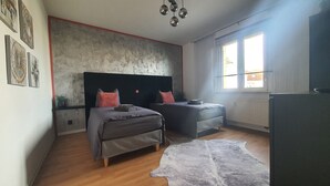 A modern bedroom with two beds, a large window, a radiator, and a ceiling light fixture.