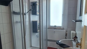 A bathroom with a glass shower enclosure, a sink, a window, and a red rug.