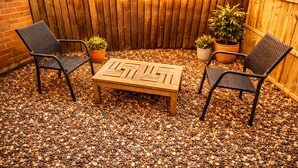 Un patio al aire libre con una mesa de madera y dos sillas de mimbre, rodeado de piedras y plantas en macetas.