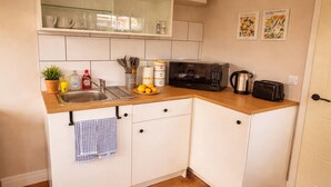 Cocina compacta equipada con microondas, tostadora y lavabo. Se observan armarios, una pequeña estantería con cristalería y una planta en maceta.