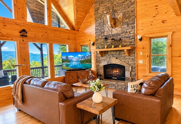 A cozy cabin interior with a stone fireplace, leather sofas, a television, and a scenic view. - Dreamy Mountain Escape, Hot Tub, Stunning mountain Views & outdoor Fireplace (Morganton)