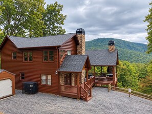 A wooden cabin with a stone chimney, a deck, and a view of a forested mountain. - Dreamy Mountain Escape, Hot Tub, Stunning mountain Views & outdoor Fireplace (Morganton)