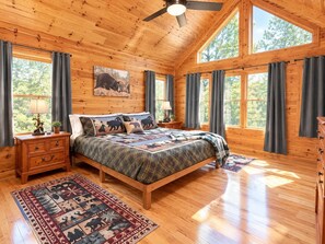 Une chambre dans une cabane en bois, avec un lit, des tables de chevet, un ventilateur de plafond et de grandes fenêtres.