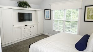 Une chambre à coucher avec un lit, un téléviseur fixé sur un meuble blanc, une fenêtre avec des stores et des œuvres d’art encadrées accrochées au mur.
