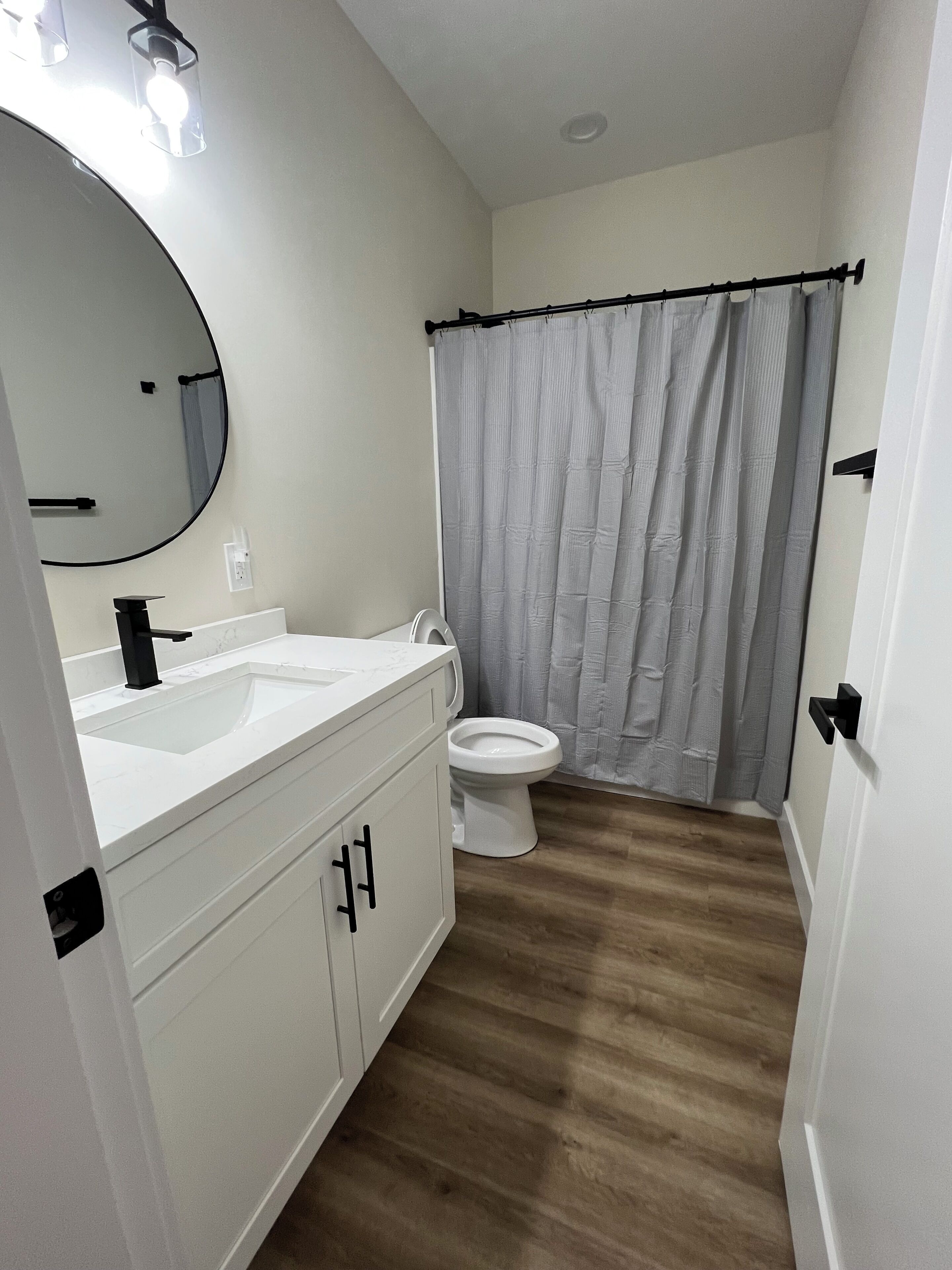 A bathroom with a white sink, mirror, and toilet, a shower curtain, and wooden flooring.