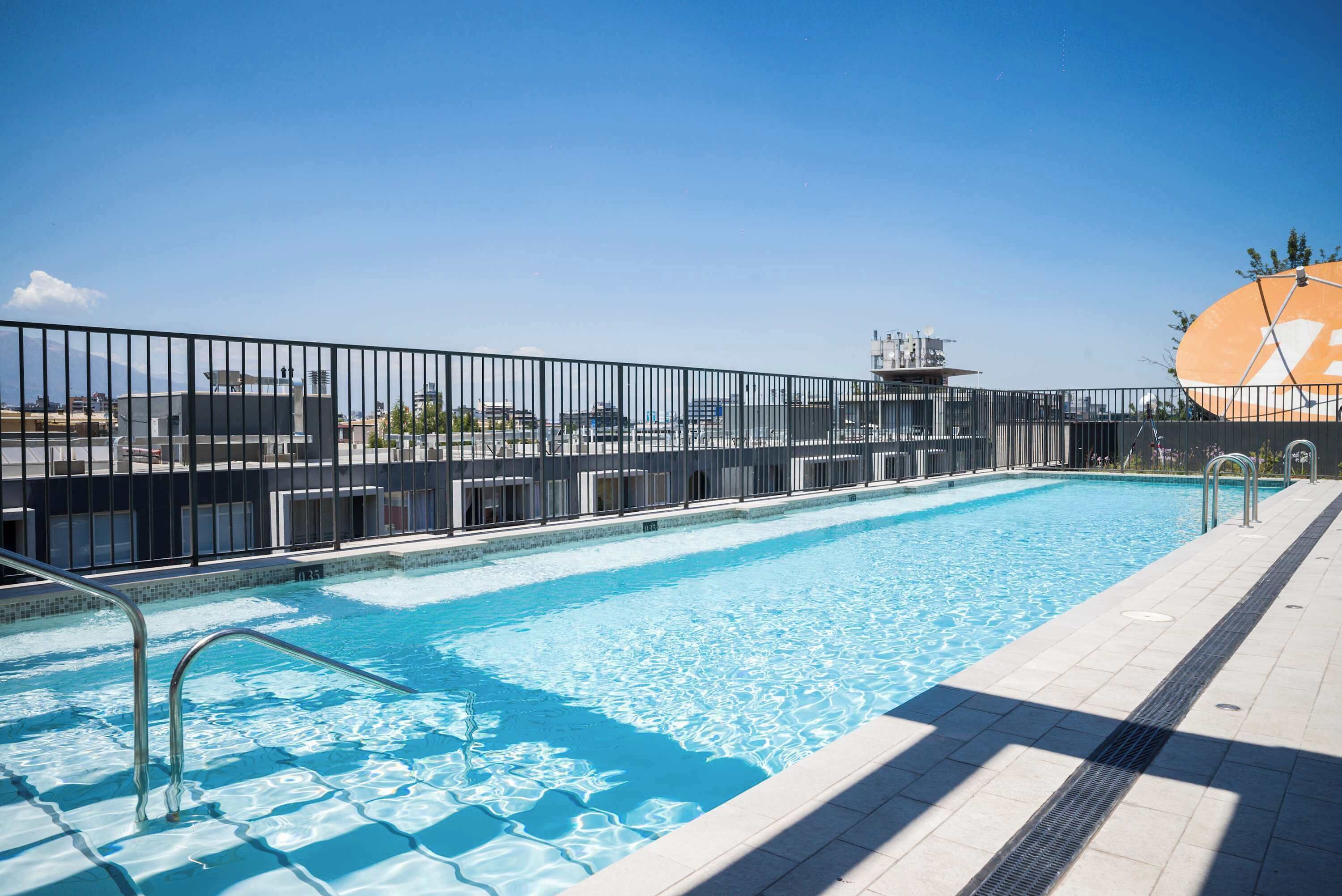 A swimming pool with a tiled deck, metal railings, and a clear blue sky in the background.