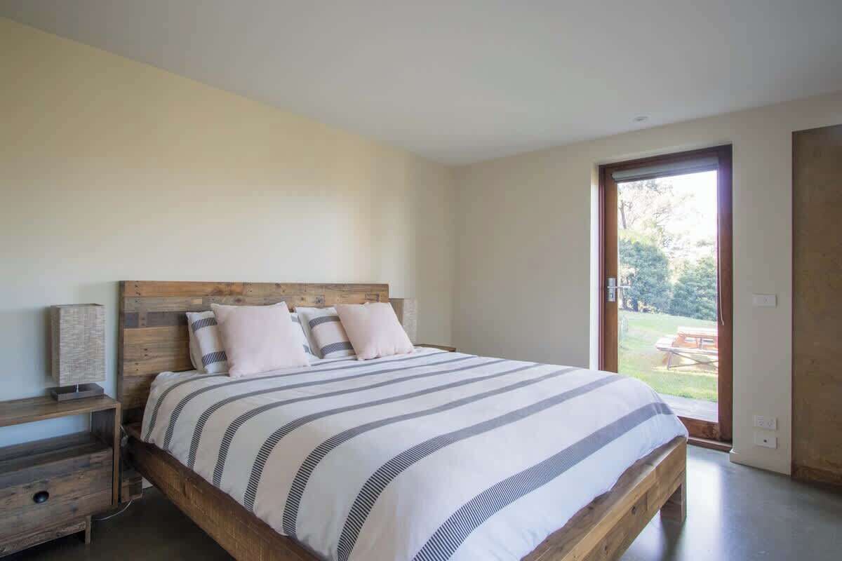 A bedroom with a wooden bed frame, a large window, and a view of the outdoors.