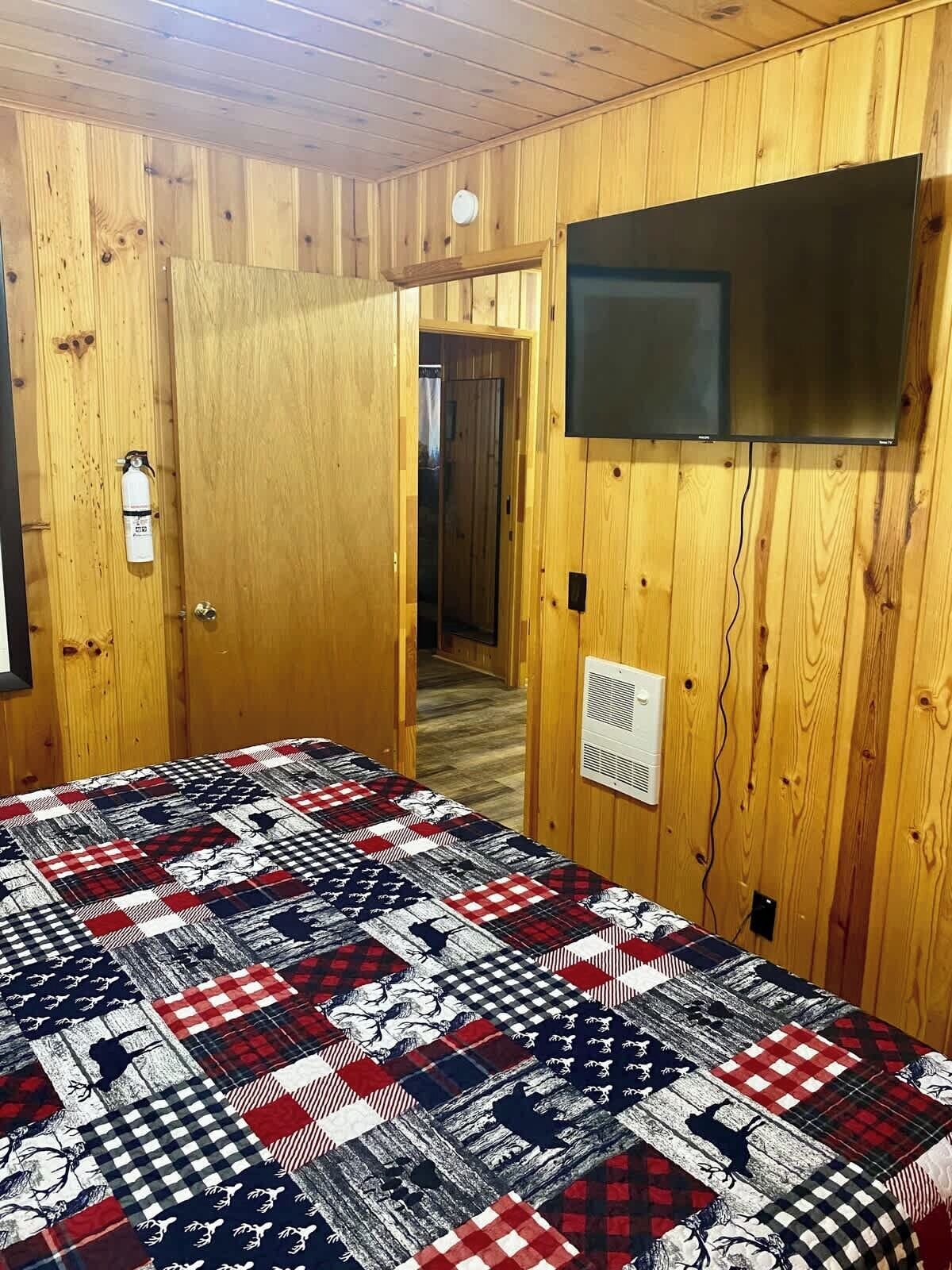 Una habitación con paredes de madera, colcha acolchada, televisor montado y una puerta con extintor.