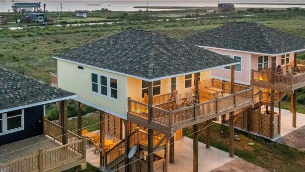 A beachfront property with multiple wooden decked houses connected by walkways, featuring outdoor seating areas and a view of the ocean. - High Noon - Hot Tub, Golf Cart, Dog Friendly! (Bolivar Peninsula)
