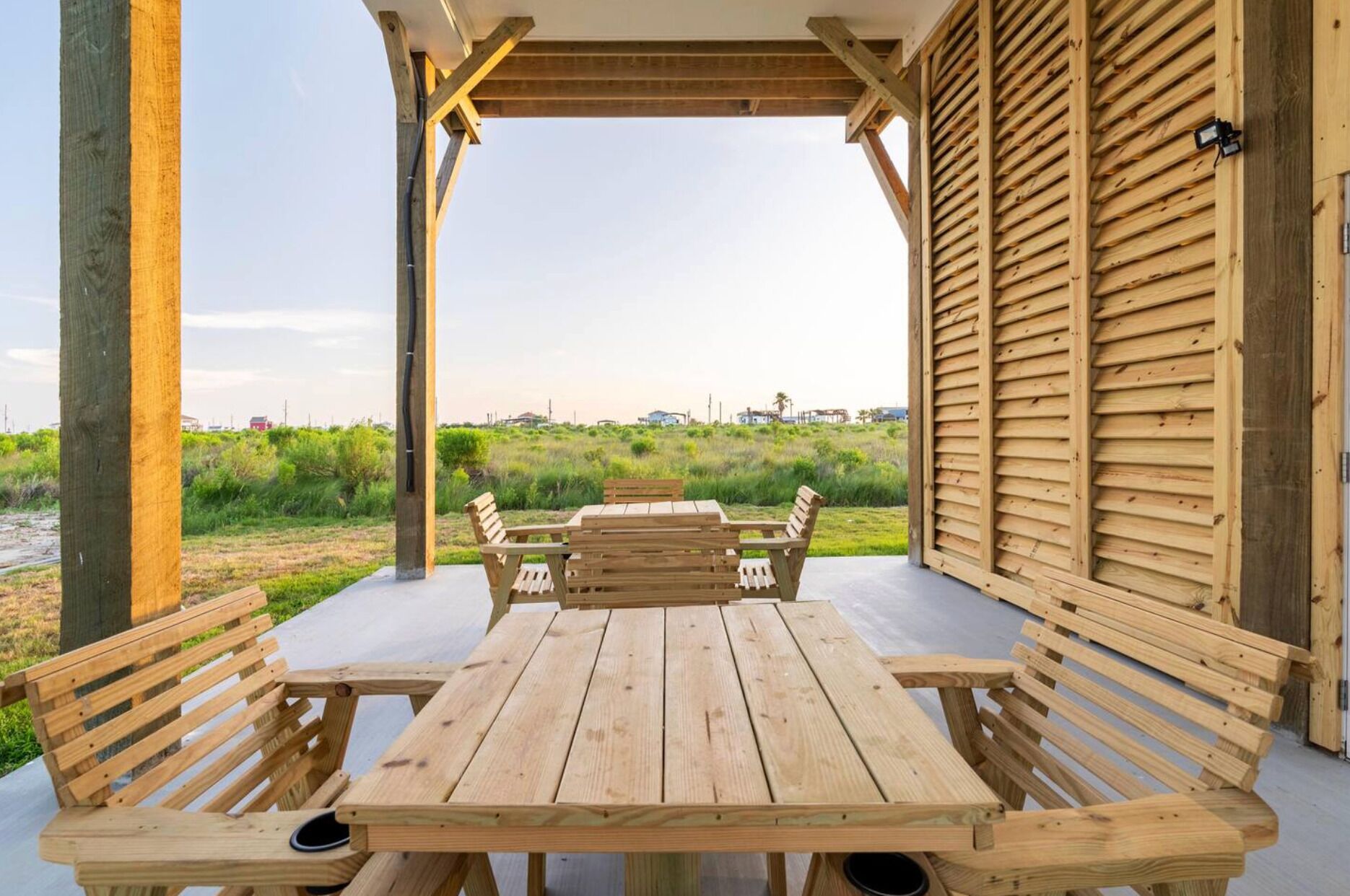 Um deck de madeira com mesa e bancos, com vista para uma área gramada e edifícios ao longe.