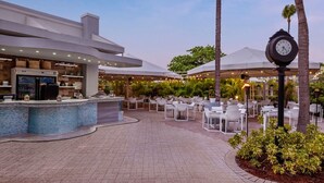 Un área de comedor al aire libre con mesas y sillas blancas, una barra con mostrador de azulejos y palmeras.