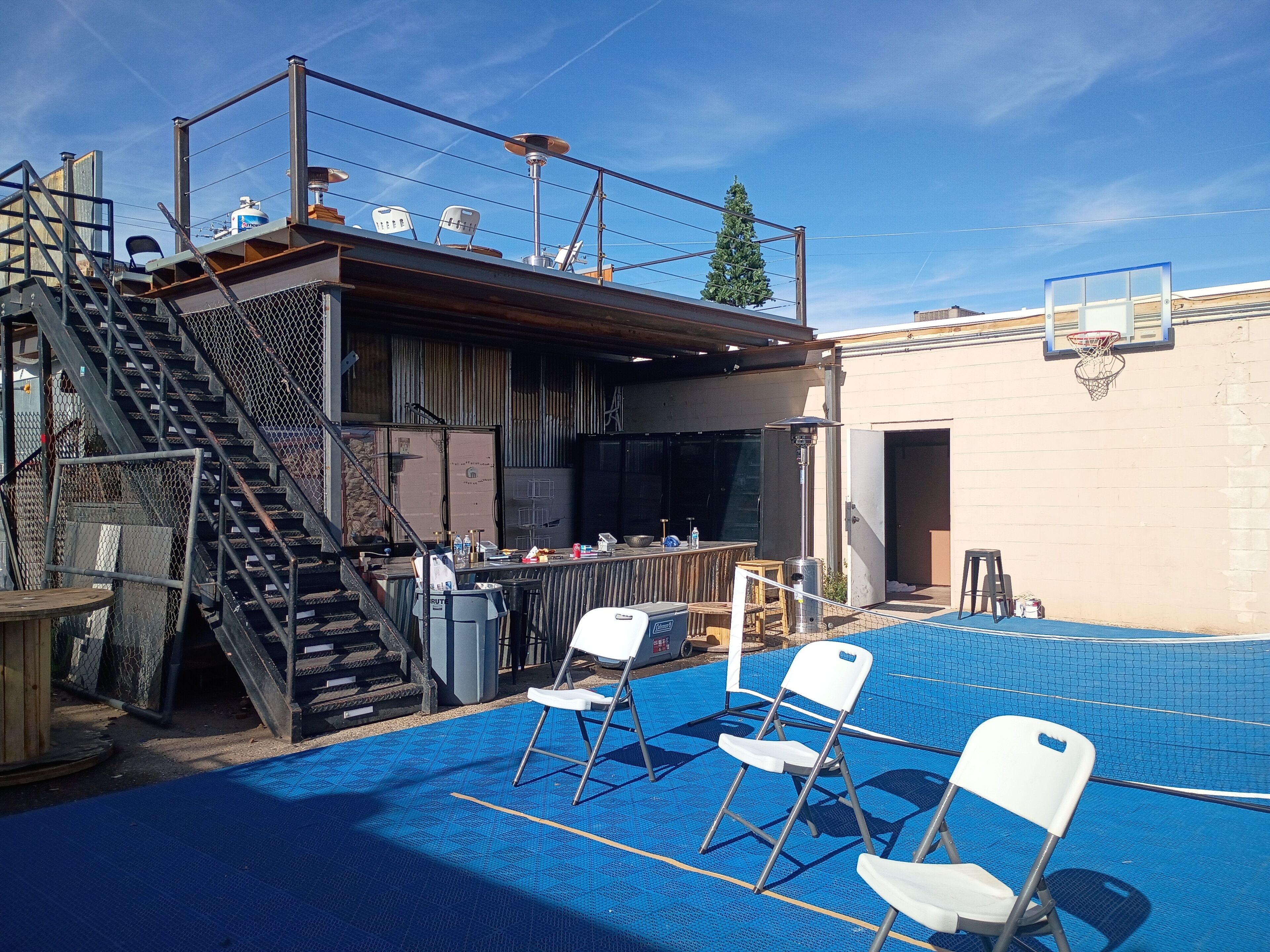 An outdoor pool area with a blue pool floor, white folding chairs, a basketball hoop, and a staircase leading to an upper deck.