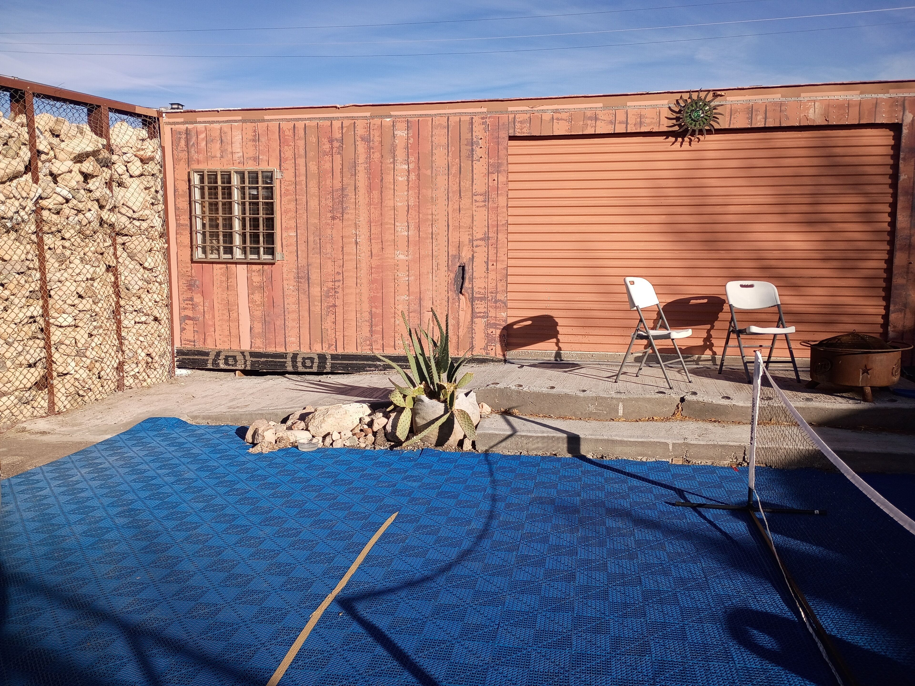 A fenced outdoor area with a blue mat, two white chairs, a cactus plant, and a metal pot.