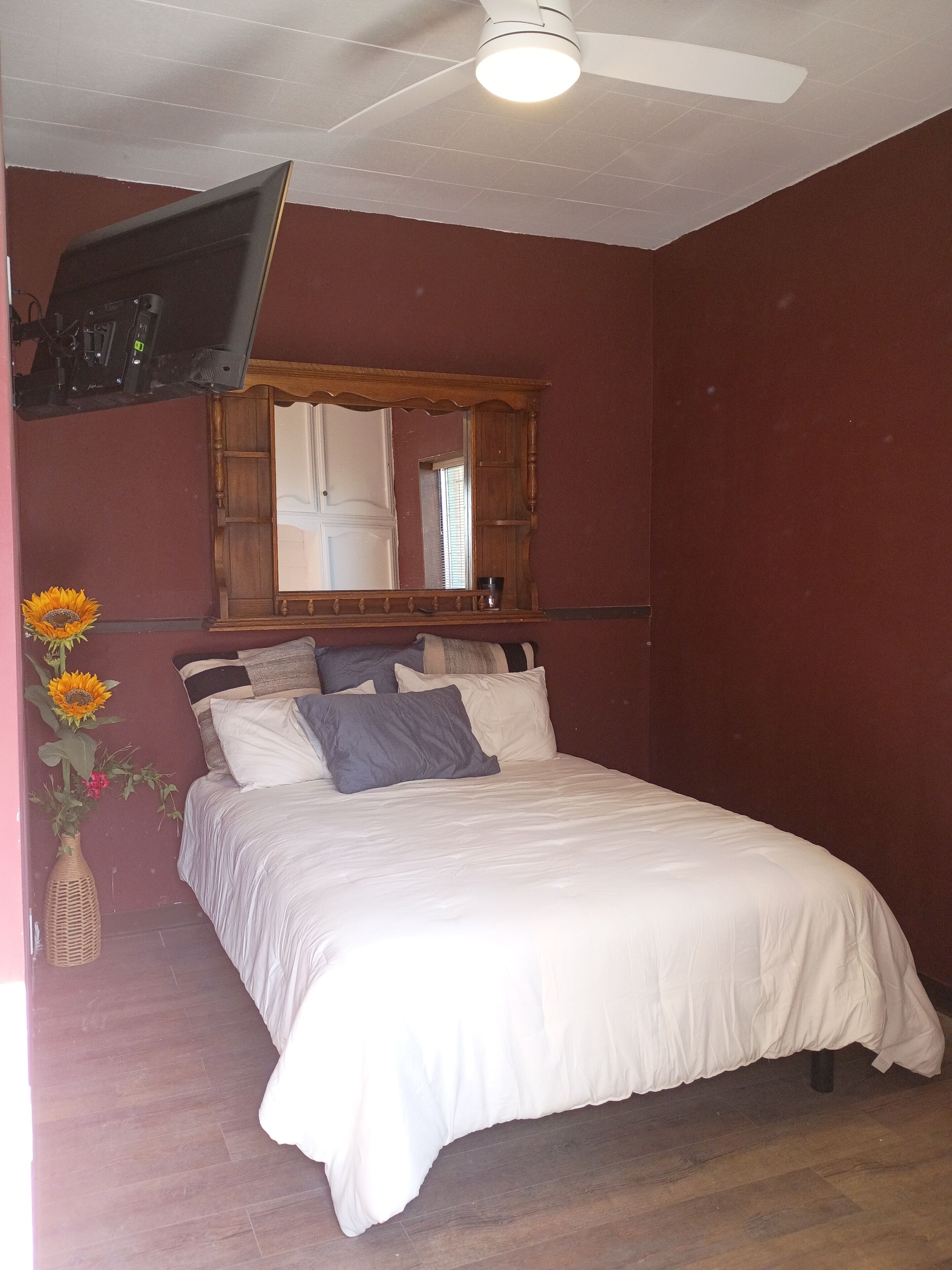 A bedroom with a bed, a television mounted on the wall, a ceiling fan, and a mirror above the bed.