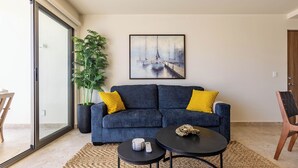 A modern living room with a dark blue sofa, yellow cushions, a round coffee table, a snake on the table, and a large potted plant. - Brisa Del Sol - Cabo Costa (Cabo San Lucas)