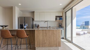 A modern kitchen with a wooden island, two bar stools, and a large window offering a view of the city. - Brisa Del Sol - Cabo Costa (Cabo San Lucas)