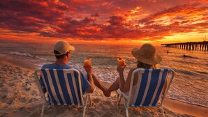 Dos personas sentadas en sillas de playa mirando al océano, sosteniendo bebidas, con un atardecer y muelle al fondo.