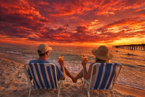 Zwei Personen sitzen auf Strandstühlen, den Ozean blickend, mit Getränken in der Hand. Im Hintergrund ist ein Sonnenuntergang und ein Steg zu sehen.
