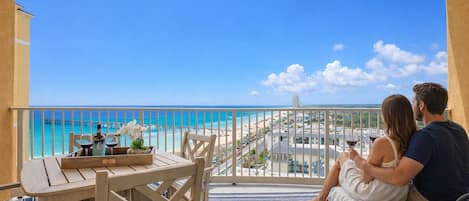 A balcony with a view of the beach and ocean, a table with a bottle and flowers, and a couple sitting on a bench.