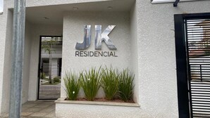 Ein Wohngebäude mit einem Schild, auf dem 'JK RESIDENCIAL' steht, einem verschlossenen Eingangsbereich und einem Pflanzbehälter mit Grünpflanzen.