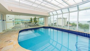 An indoor swimming pool with a glass ceiling, clear blue water, and a view of the sea. - Apto c Piscina, Garagem e Academia a 180m da Praia (Guarujá)