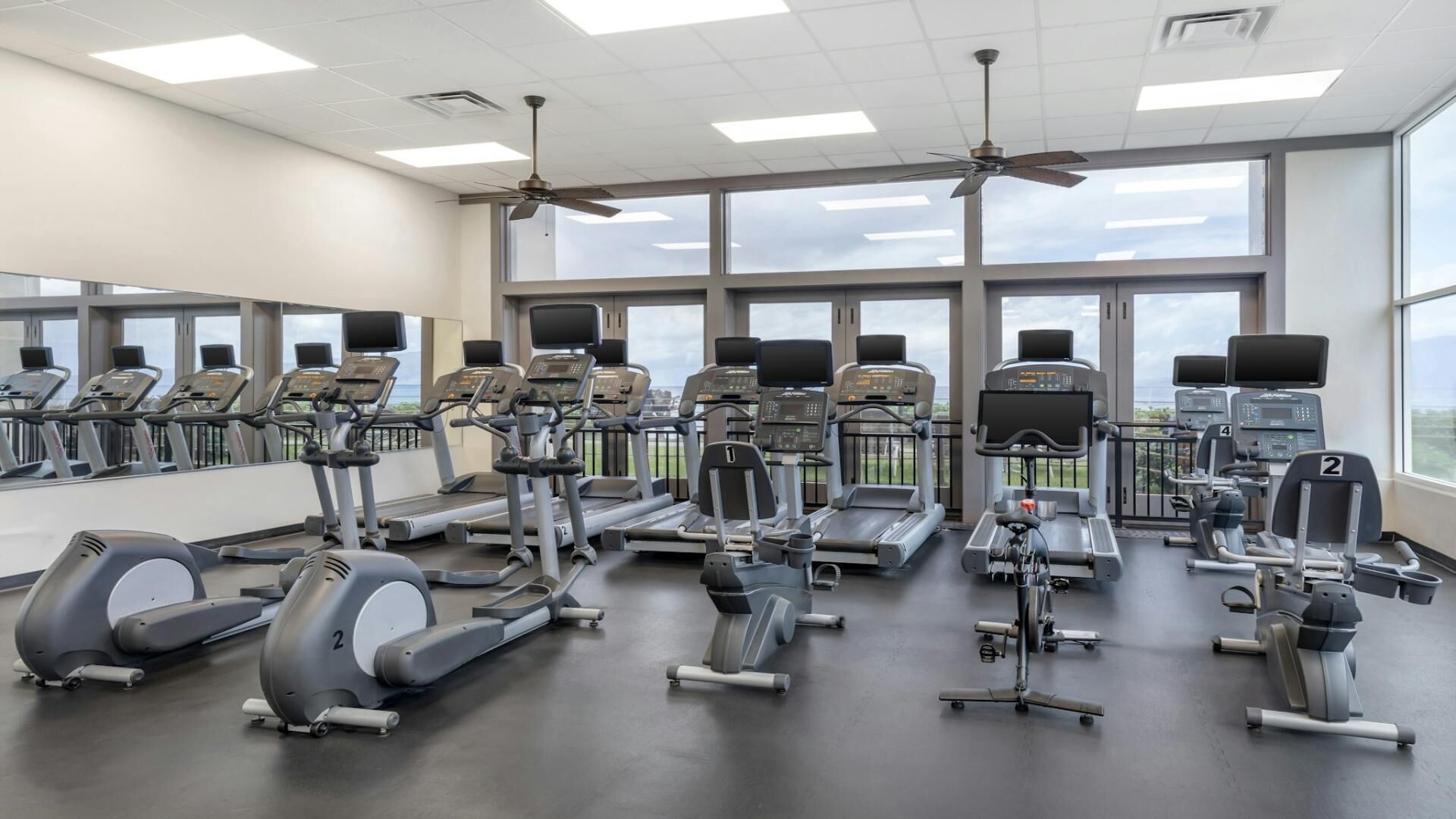 Et fitnesscenter med flere løbebånd, crosstrainere og spinningcykler, store vinduer og en loftsvifte.
