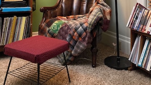 Un coin lecture confortable avec un fauteuil en cuir, un pouf rouge et une lampe sur pied. Une bibliothèque remplie de livres se trouve à gauche, et un tableau représentant un oiseau est accroché au mur.