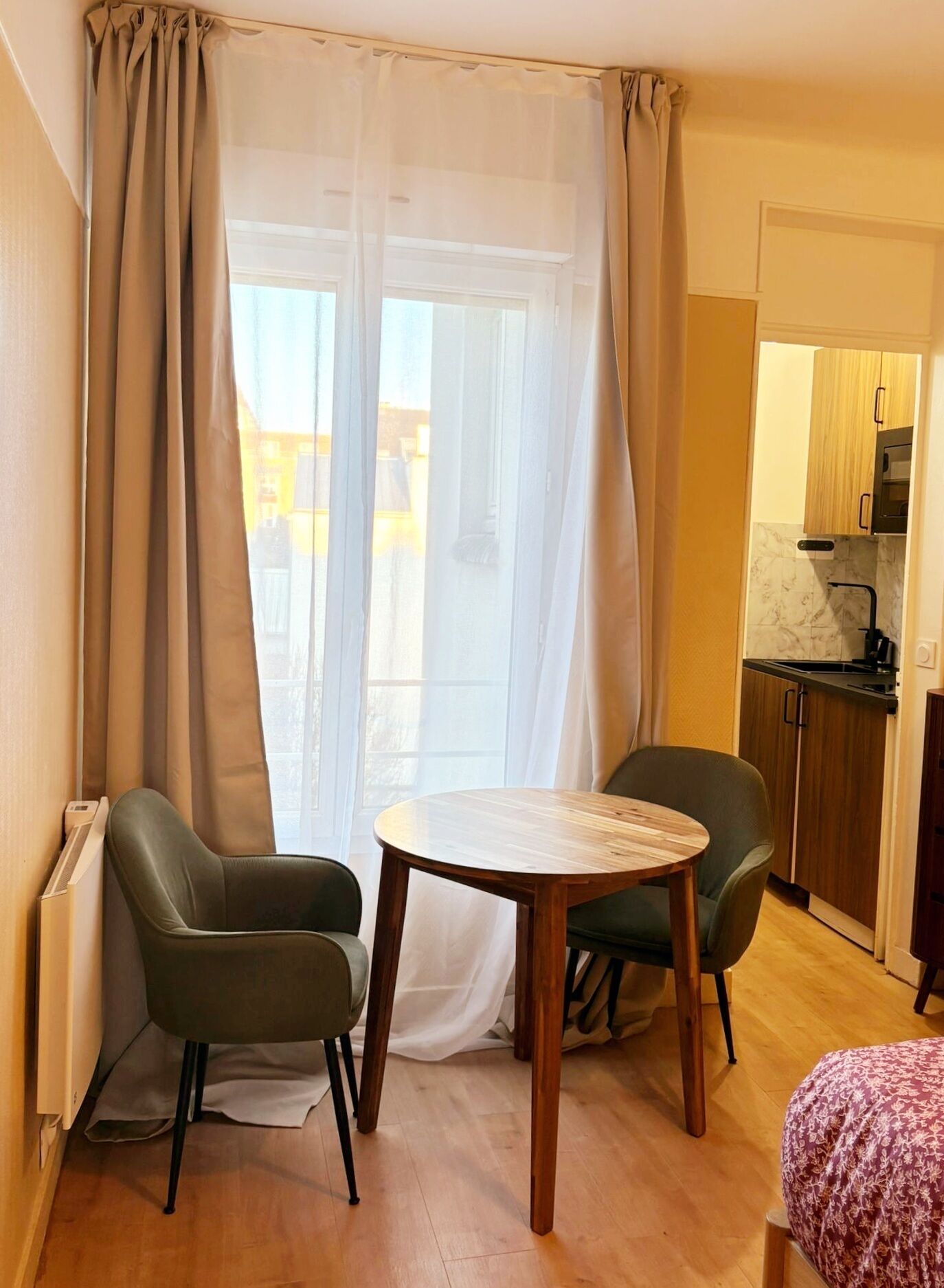 Ein Zimmer mit einem runden Holztisch, zwei Stühlen, einem Heizkörper, einem Fenster mit durchscheinenden Vorhängen sowie einer Küchenzeile mit Holzschränken.