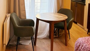 Una habitación con una mesa redonda de madera, dos sillas, un radiador, una ventana con cortinas translúcidas y una zona de cocina con armarios de madera.
