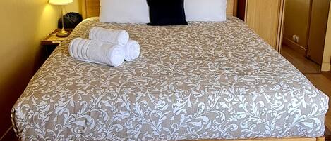 A neatly made bed with a patterned bedspread, a black pillow, and rolled towels on the side.
