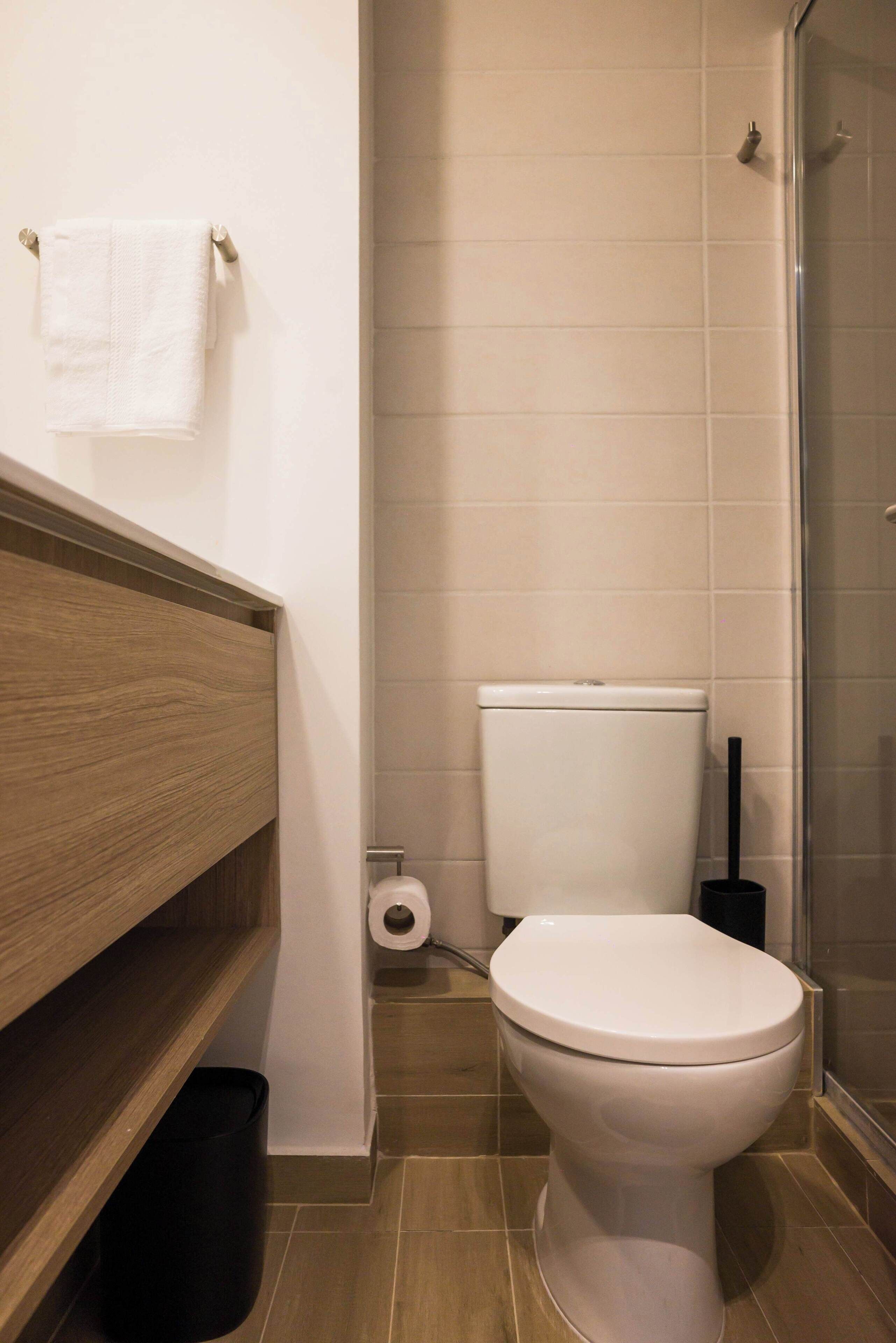 Une salle de bain moderne avec des toilettes, une étagère en bois, une poubelle noire et une cabine de douche en verre.