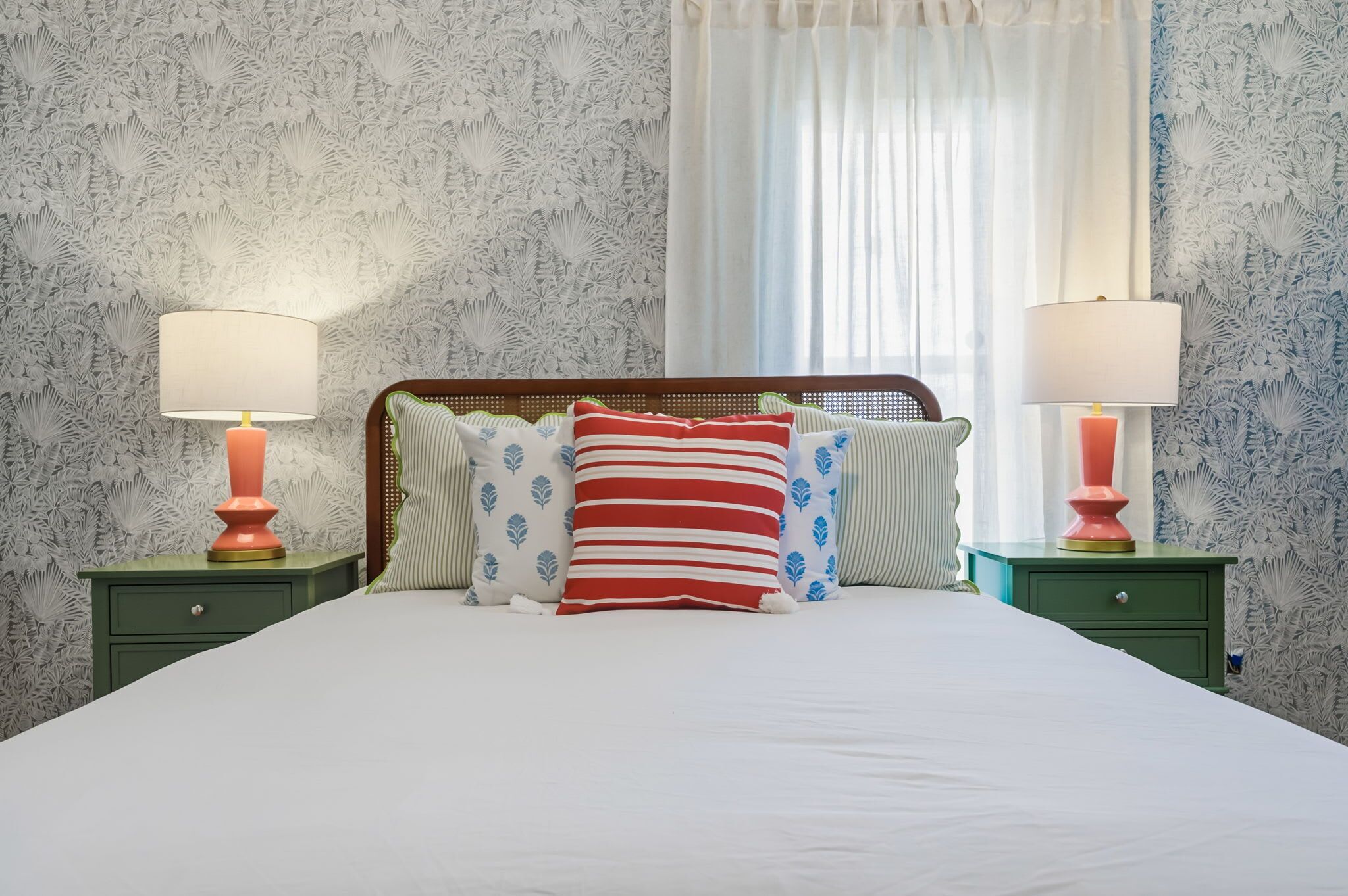 A neatly made bed with a white comforter and striped pillows, flanked by two green nightstands with lamps, against a wall with a leafy pattern.