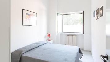 Un dormitori amb un llit, una finestra, un radiador i una cambrera amb una làmpada.