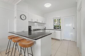 A modern kitchen with white cabinets, a black countertop, and wooden stools. - Villa 03 Tangalooma Donegal (Tangalooma)