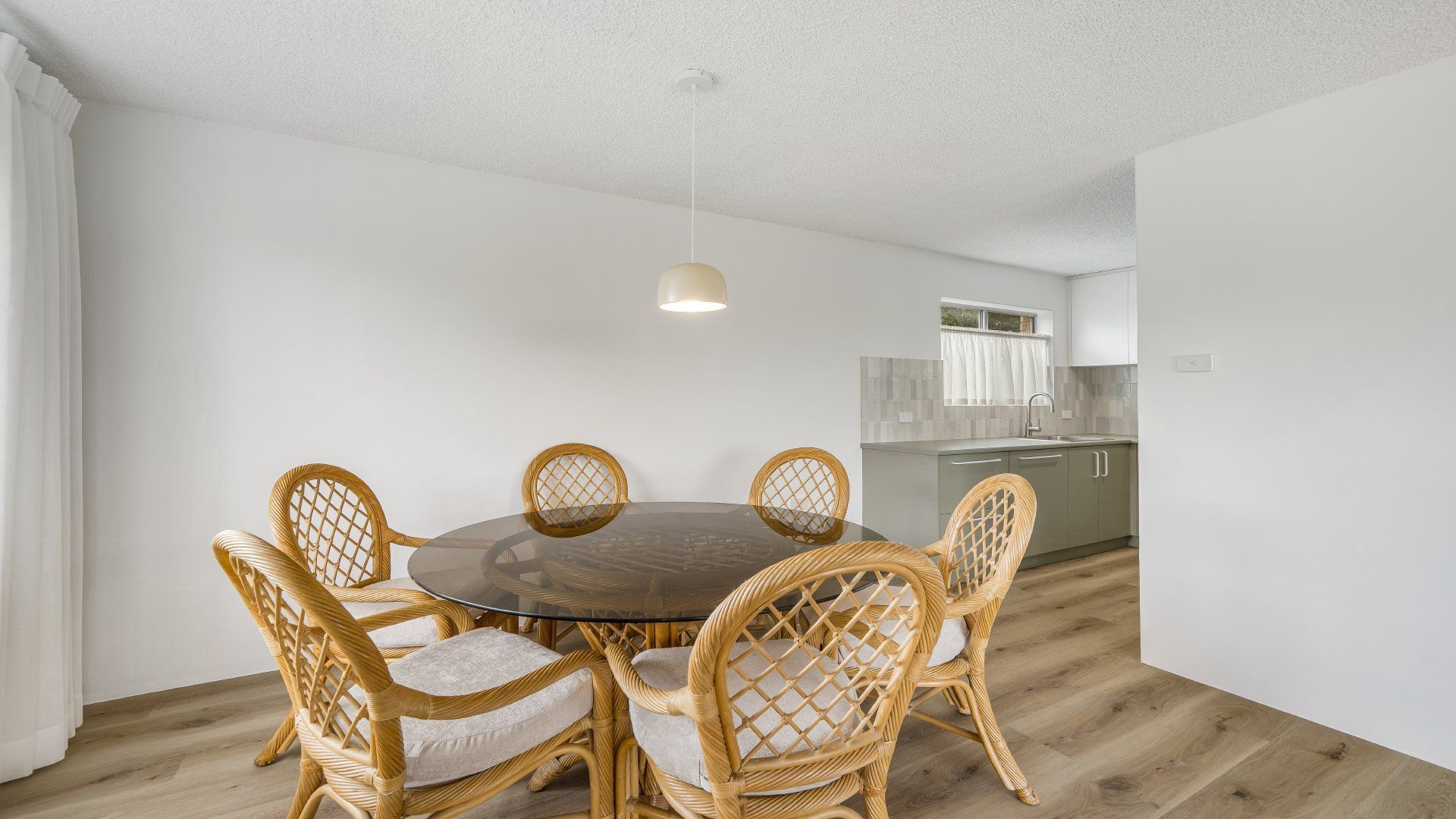 A dining area with a round table and wicker chairs, a kitchen in the background, and a white ceiling.