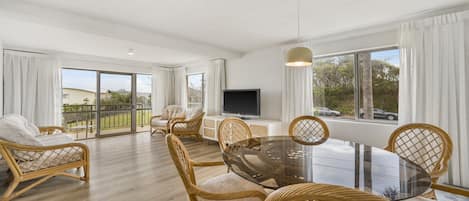 A spacious living area with a glass-top dining table surrounded by wicker chairs, a flat-screen TV on a white cabinet, and large windows offering a view of the outdoors.