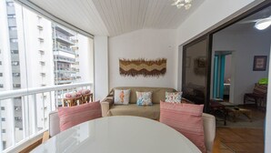 A modern living room with a large window, a round table, and a sofa with patterned cushions. - Apto c Piscina Garagem e Academia a 180m da Praia (Guarujá)