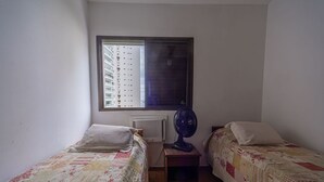 A small, single-room with two beds, a window with blinds, a standing fan, and a radiator. - Apto c Piscina Garagem e Academia a 180m da Praia (Guarujá)
