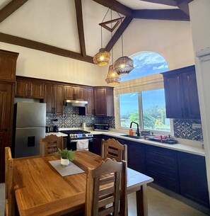 Une cuisine avec des meubles en bois, un réfrigérateur en acier inoxydable, une cuisinière et une table à manger avec des chaises. On y trouve également des luminaires suspendus et une fenêtre avec vue.