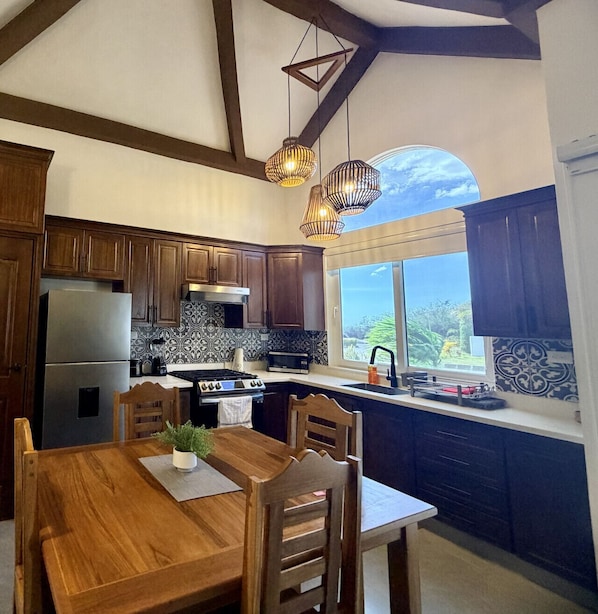 A kitchen with wooden cabinets, a stainless steel refrigerator, a stove, and a dining table with chairs. There are hanging pendant lights and a window with a view.