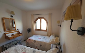 Un dormitorio con cama, mesita de noche, espejo, ventana con cortinas y lámpara de pared.