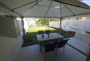 Un patio exterior con una mesa y sillas bajo una marquesina, rodeado por un jardín vallado con césped y plantas en macetas.
