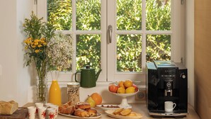 Frühstücksecke mit Kaffeemaschine, Früchten, Gebäck und einem Blumenstrauß an einem Fenster mit Blick auf Grünflächen.