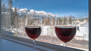 Two wine glasses on a windowsill with a view of snow-capped mountains and a town in the background. - Spacious Dual-Unit Stay | 2 Addresses | Ideal for Large Groups (Canmore)