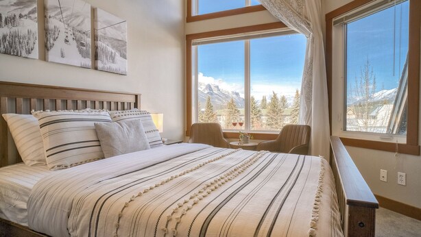 A bedroom with a bed, a nightstand, a chair, and a window with a mountain view. - Spacious Dual-Unit Stay | 2 Addresses | Ideal for Large Groups (Canmore)